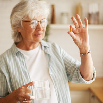 Beautiful Mature Sixty Year Old Female In Stylish Eyeglasses Holding Mug And Omega 3 Supplement Capsule, Going To Take Vitamin After Meal. Senior Gray Haired Woman Taking Fish Oil Pill With Water