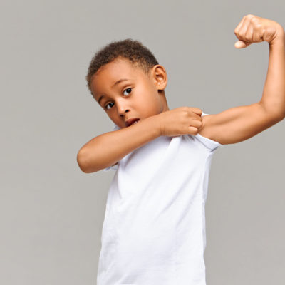 Look At My Bicep. Picture Of Funny Afro American In Casual White T Shirt Posing Isolated At Gray Studio Wall Pulling Up Sleeve, Showing His Tensed Arm Muscle. Childhood, Fitness And Sports Concept