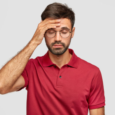 Overworked Cuacasian Male Feels Terrible Headache After Sleepless Night, Keeps Hand On Forehead, Shut Eyes, Dressed In Casual Red T Shirt, Stands Against White Background. People And Tiredness