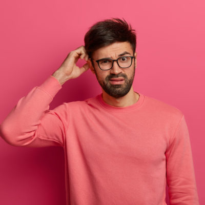 Serious Bearded Young Man Scratches Head, Thinks Deeply About How To Solve Problem, Makes Decision, Wears Optical Glasses, Poses Indoor Against Rosy Background, Makes Up Plan, Has Bad Memory