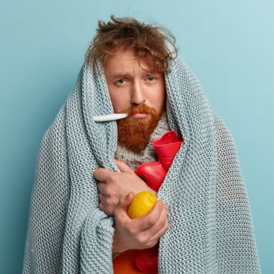 Redhead Young Man With Bristle Fights Against Flu And Fever, Holds Lemon To Enrich Vitamins, Has Thermometer In Mouth, Trembles Under Blanket, Isolated Over Blue Background. Health Problems.