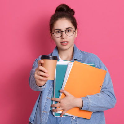 Student Girl With Dark Hair Offers Takeaway Coffee, Holding Paper Folder In Hands, Standing Isolated Over Pink Background, Lady Has Coffee Break Between Lectures.