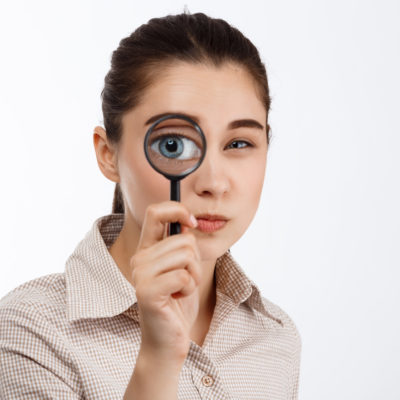 Young Beautiful Brunette Girl Looking With Mistrust At Camera Through Magnifier Over White Background.